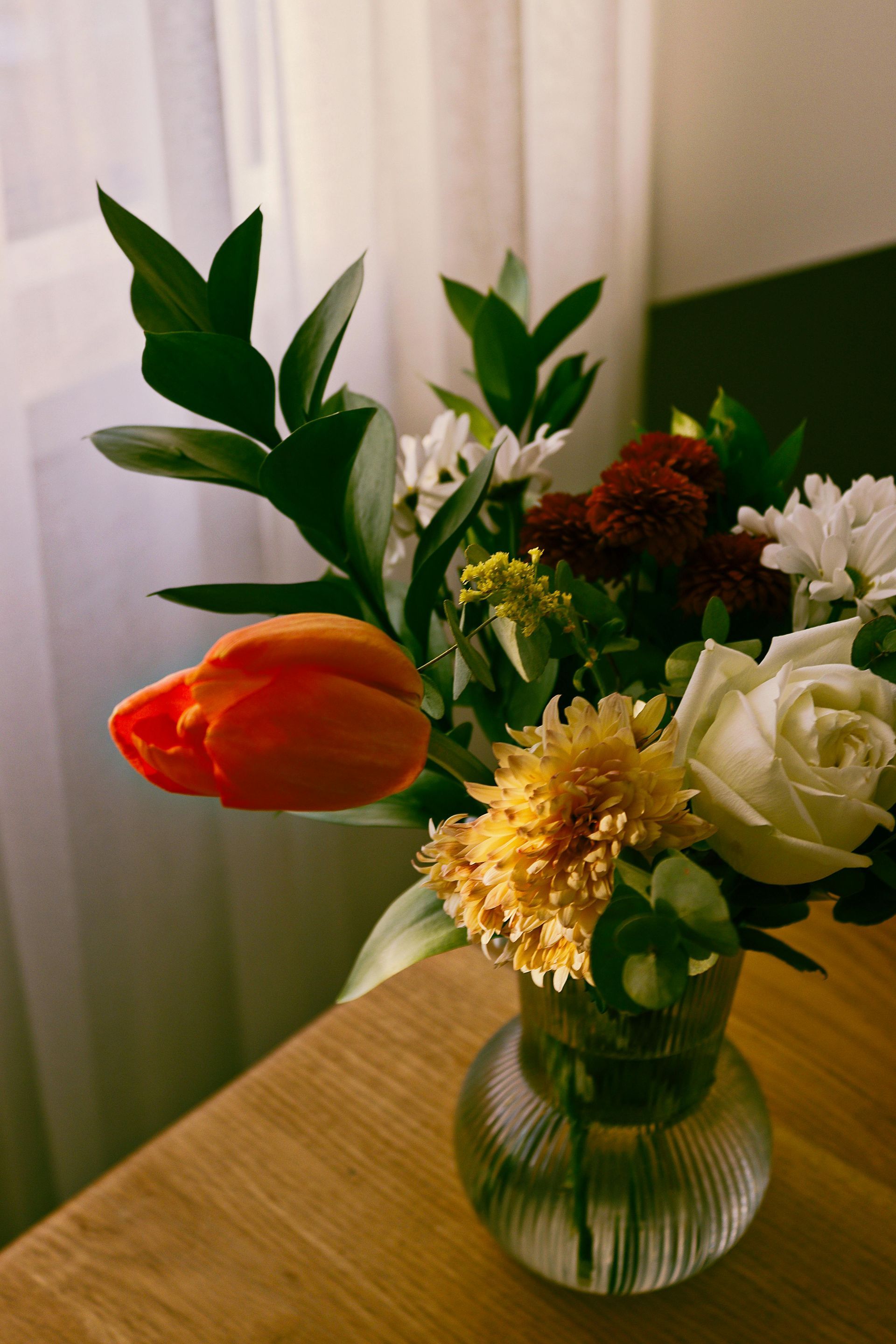 Un vase de fleurs variées, dont une tulipe rouge, posé sur une table en bois près d'une fenêtre.