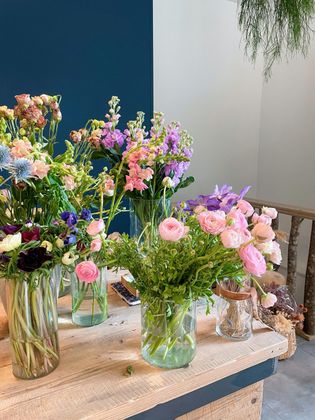 Des fleurs aux tons pastel assortis, présentées dans des vases en verre, sont disposées sur une table devant un mur bleu et blanc.