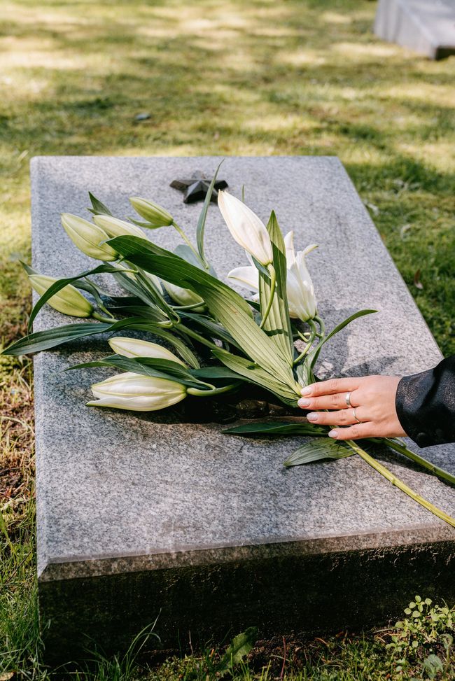Un bouquet de lys blancs et de verdure sur une tombe de pierre dans un cimetière herbeux ; une main repose à côté.