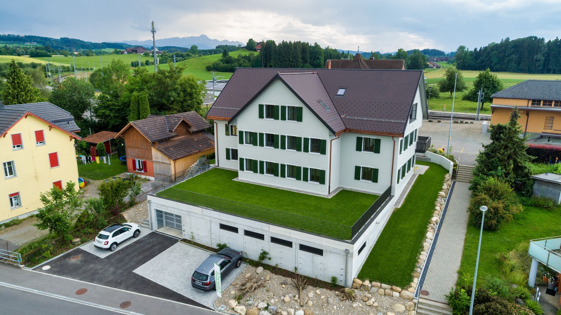 Luftaufnahme eines weißen Hauses mit braunem Dach neben Feldern und Nachbarhäusern in einem ländlichen Dorf.