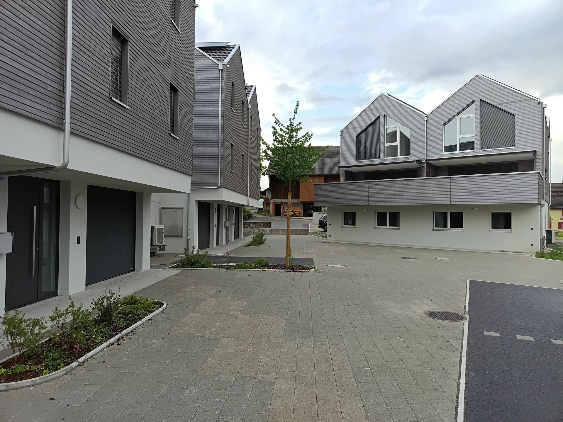 Moderner Apartmenthof mit grauen Gebäuden, gepflasterter Terrasse und einem kleinen Baum unter bewölktem Himmel