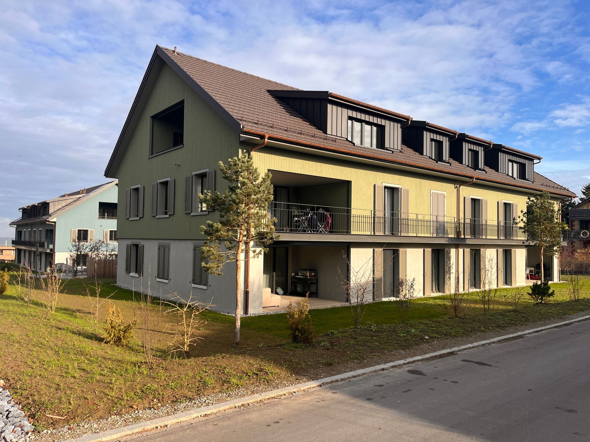 Modernes beigefarbenes Mehrfamilienhaus mit Balkonen an einer ruhigen Straße unter einem teilweise bewölkten Himmel