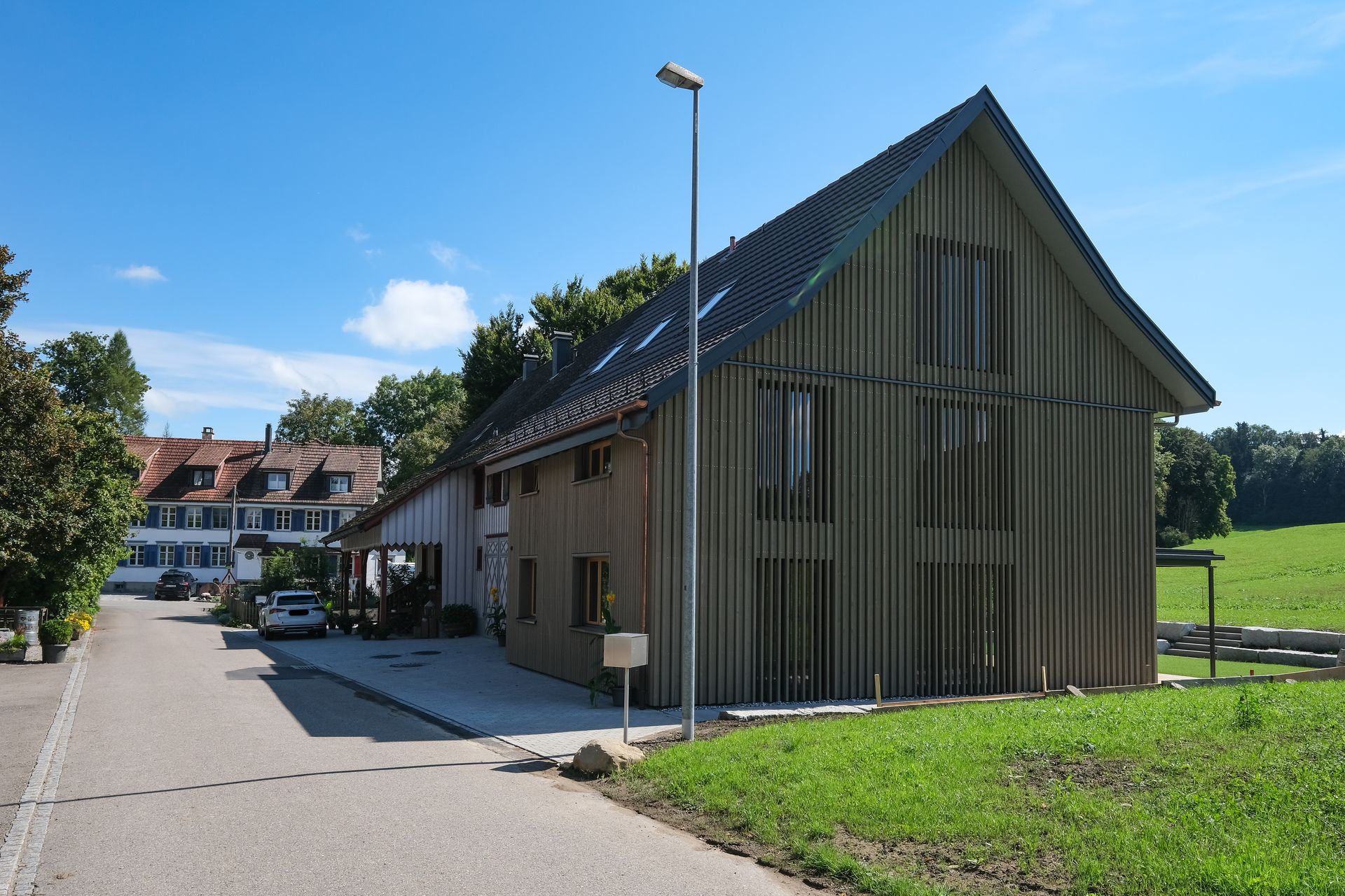 Modernes Gebäude mit schrägem Dach neben einer Straße, umgeben von Häusern, Bäumen und grünen Feldern unter blauem Himmel