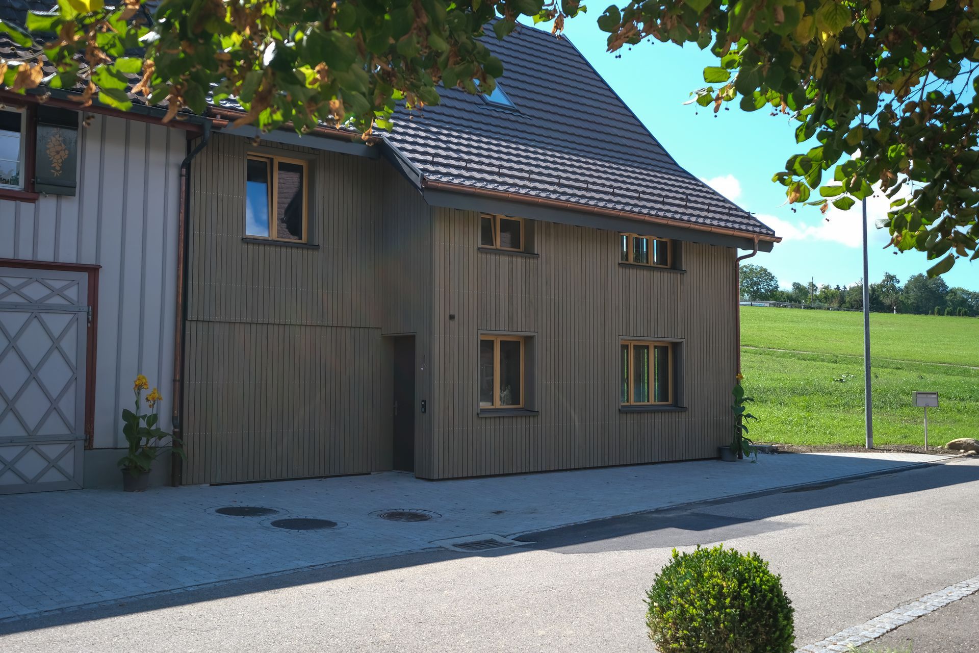 Holzhaus mit Ziegeldach neben einer gepflasterten Straße, im Hintergrund grüne Felder.