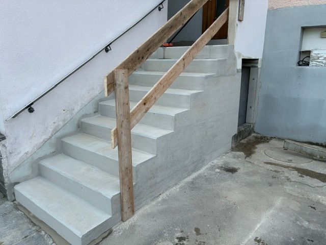 Ein weiss getünchtes Haus mit grünem Rand in einem Hofparkplatz, mit Autos, einem blauen Parkschild und kahlen Bäumen.