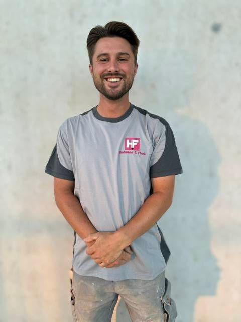 Eine lächelnde Person steht vor einer hellen Wand und trägt ein graues Hemd mit einem rosa Logo.