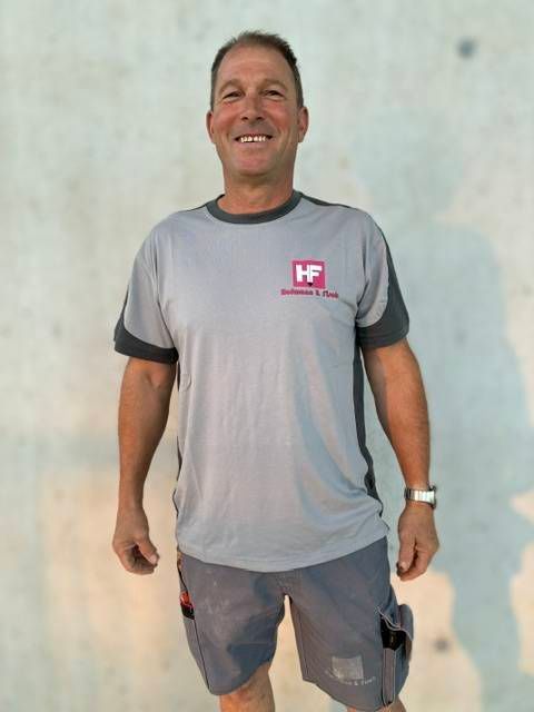 Ein lächelnder Mann steht an einer schlichten Wand; er trägt ein graues T-Shirt und Shorts.