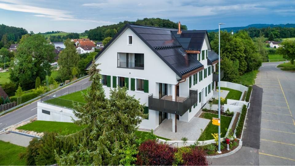 Modernes weisses Haus mit dunklem Dach, Balkonen und angelegtem Garten neben einem leeren Parkplatz