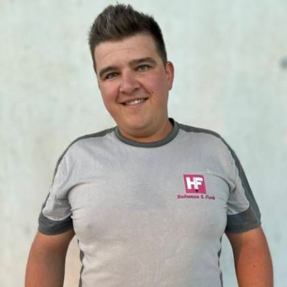 Eine Person in einem grauen T-Shirt und dunkler Hose steht vor einer hellen Wand und lächelt in die Kamera.