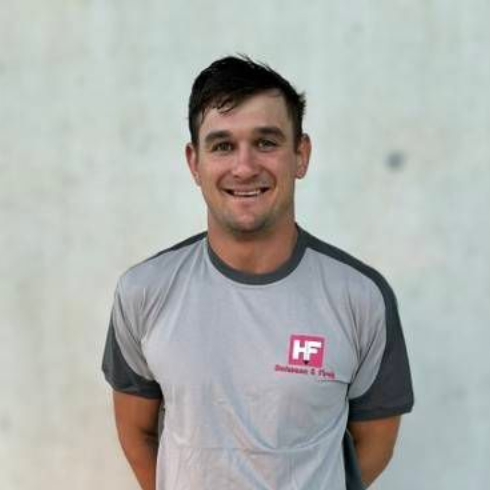 Ein Mann, der in einem grauen T-Shirt und Shorts an einer hellen Wand steht, lächelt mit den Händen hinter dem Rücken.
