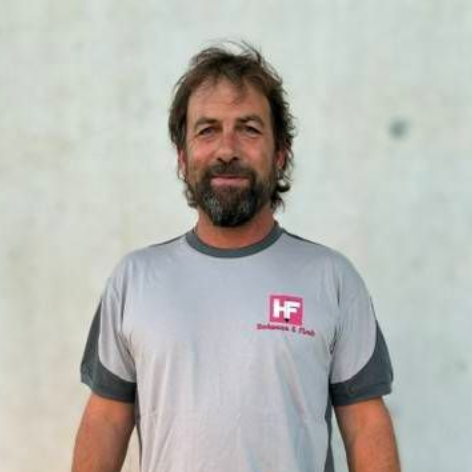Ein Mann, der an einer hellen Wand steht, ein graues T-Shirt und hellbraune Shorts trägt, schaut in die Kamera.