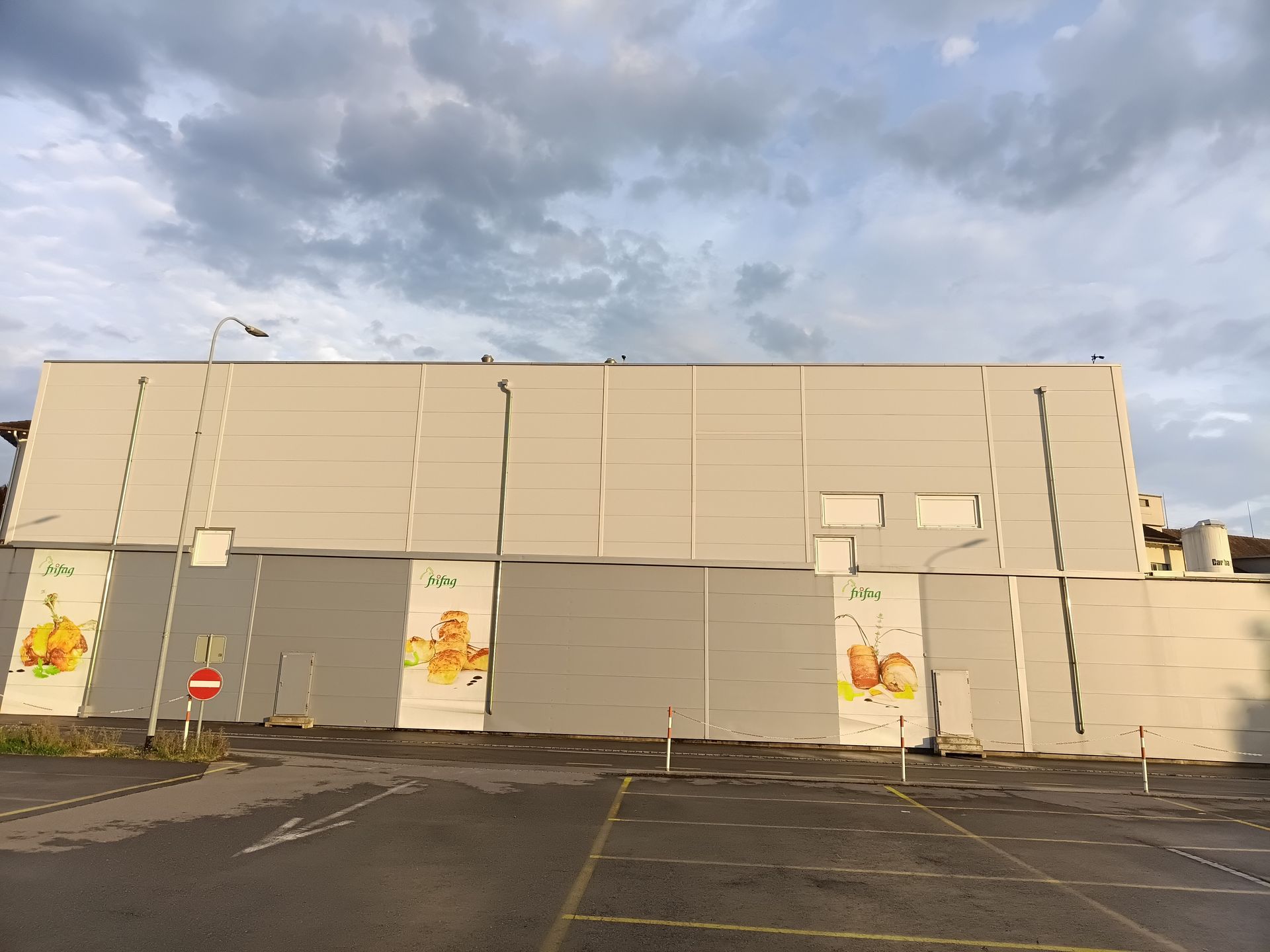 Großes beigefarbenes Industriegebäude mit grauer unterer Fassade und bewölktem Himmel über einem leeren Parkplatz