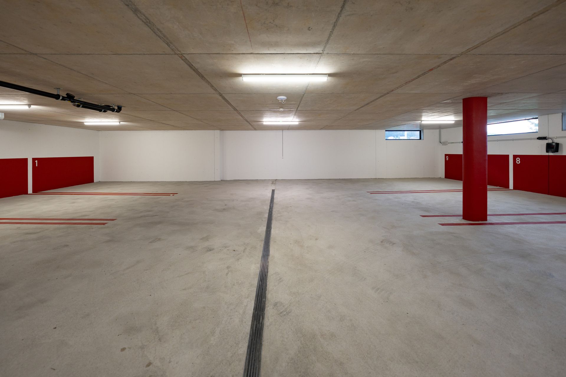 Leere Tiefgarage mit weißen Wänden, Betonboden, roten Säulen und Leuchtstoffröhren an der Decke.