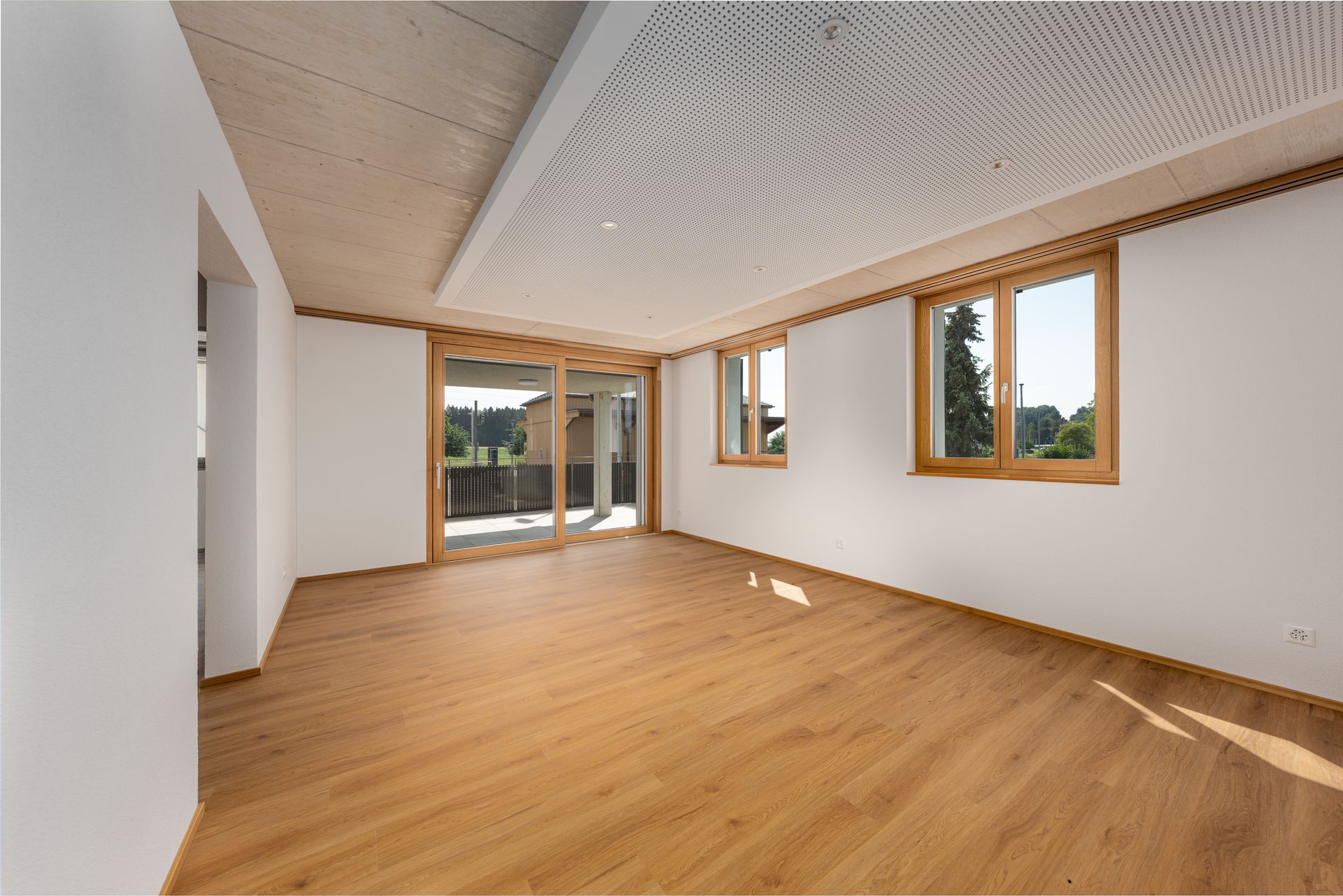 Leeres, modernes Zimmer mit Holzboden, weißen Wänden und großen Fenstern, die auf einen Balkon führen.