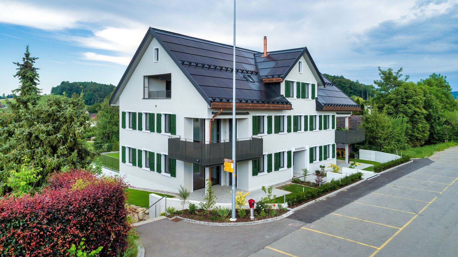 Weißes Haus mit grünen Fensterläden an einer Straße, umgeben von Bäumen und Sträuchern unter einem bewölkten Himmel