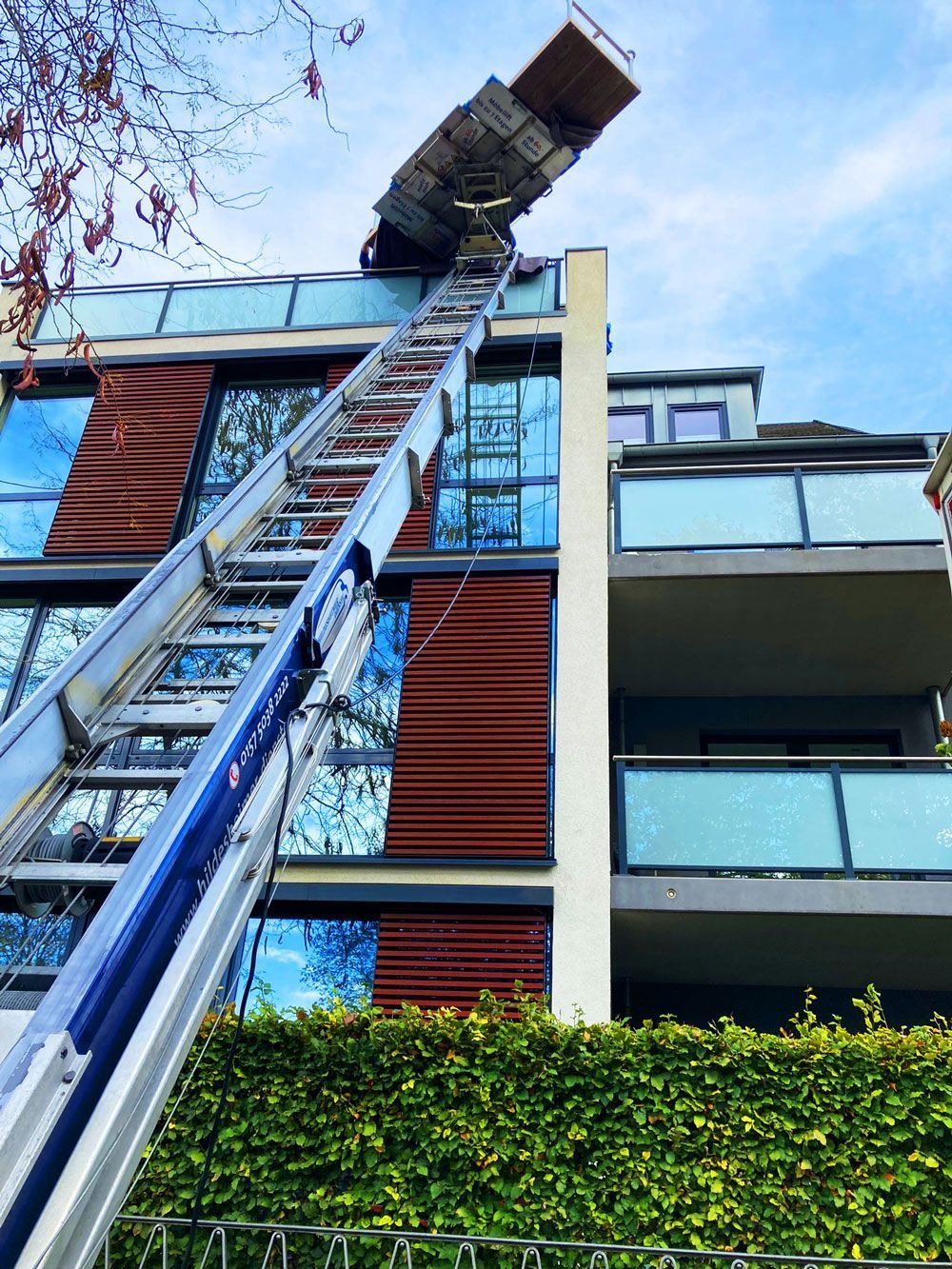Ein Möbellift im Einsatz an einem modernen Wohngebäude. Über eine lange Schiene werden Möbel oder Baumaterialien sicher in die oberen Etagen transportiert. Jetzt Umzugslift mieten in Hildesheim für einen schnellen und bequemen Umzug ohne Treppensteigen!