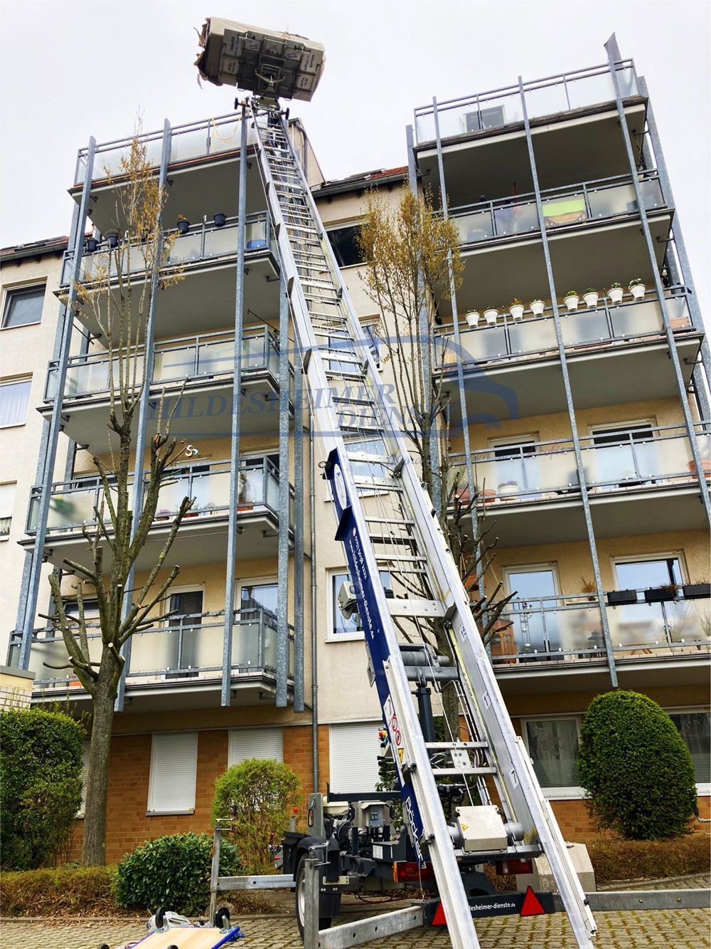 Ein Mehrfamilienhaus hat einen Möbellift in Hildesheim gemietet. Dieser ist an dem Mehrfamilienhaus aufgebaut. Die lange, schräg aufgestellte Leiterkonstruktion reicht bis zum oberen Stockwerk, wo eine Plattform mit Umzugsgut beladen ist. Der Lift ist auf einem Anhänger montiert, der auf der Straße vor dem Gebäude steht. Die Balkone des Hauses sind mit Glasgeländern versehen, und einige Bäume stehen vor dem Gebäude.