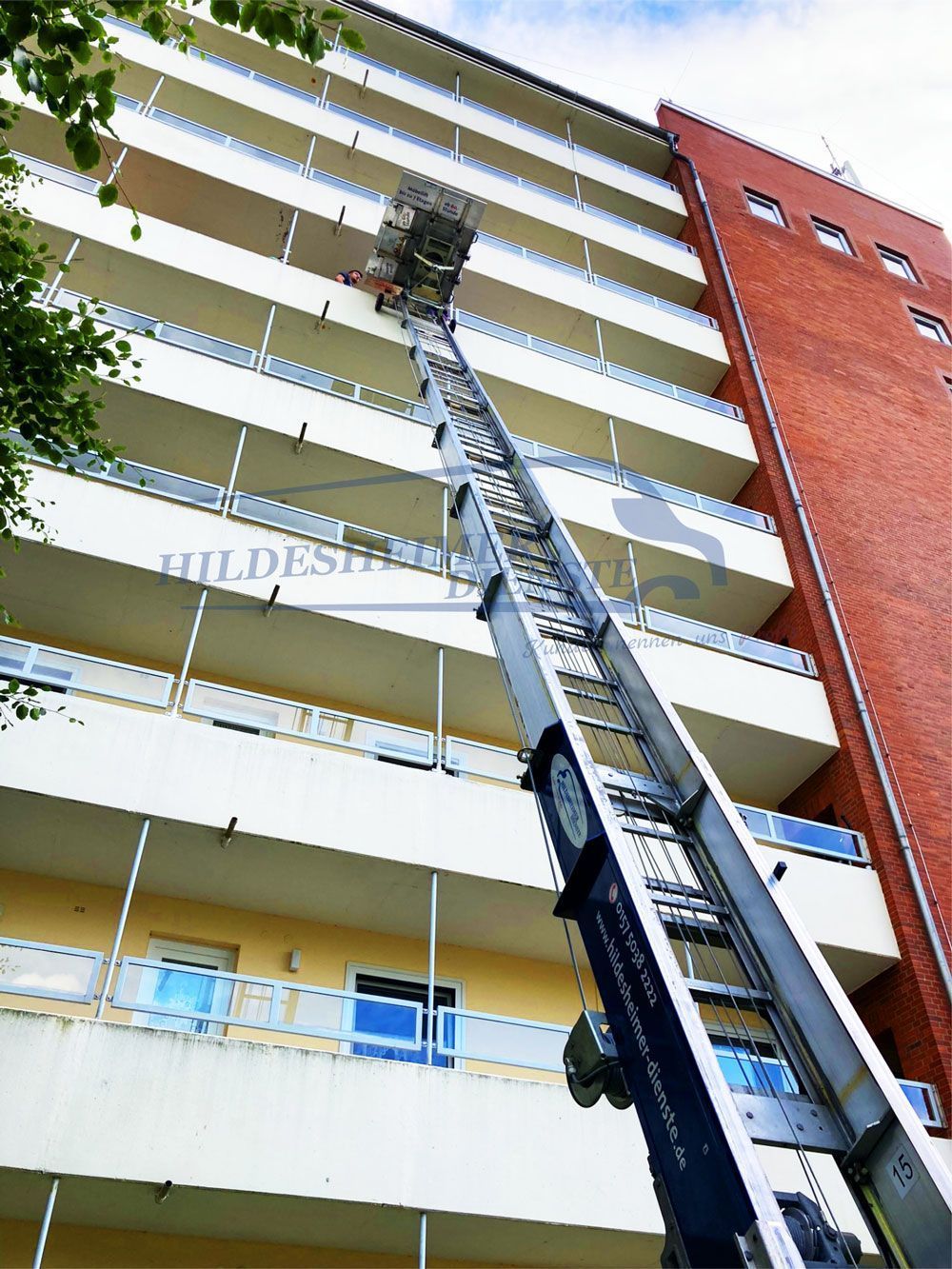 Ein Möbellift wurde in Hildesheim gemietet und an einem hohen Wohngebäude positioniert. Die lange, schräge Hebebühne erstreckt sich bis in die obersten Etagen, wo eine Plattform mit Umzugsgut beladen wird. Der Lift ist auf einem Fahrzeug montiert und steht auf dem Boden vor dem Gebäude. Die Fassade kombiniert gelbe und rote Elemente, während die Balkone mit weißen Geländern versehen sind. Im Vordergrund sind einige Äste von Bäumen sichtbar.