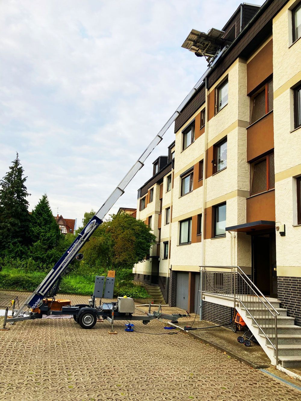 Ein Umzugslift kann in Hildesheim gemietet werden und ist hier an einem mehrstöckigen Wohnhaus aufgestellt. Die schräg verlaufende Hebebühne transportiert eine Plattform mit Umzugsgut bis ins oberste Stockwerk. Der Lift ist auf einem Anhänger montiert und steht auf einem gepflasterten Hof vor dem Gebäude. Eine Treppe mit Geländer führt zum Hauseingang, während im Hintergrund Bäume und weitere Gebäude zu sehen sind.