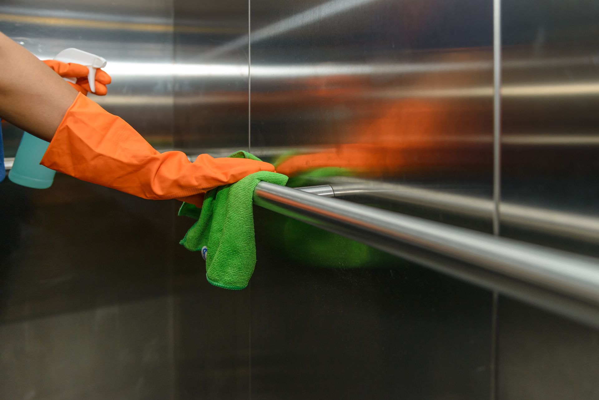 Mains portant des gants oranges nettoyant une rambarde d'ascenseur avec un chiffon vert