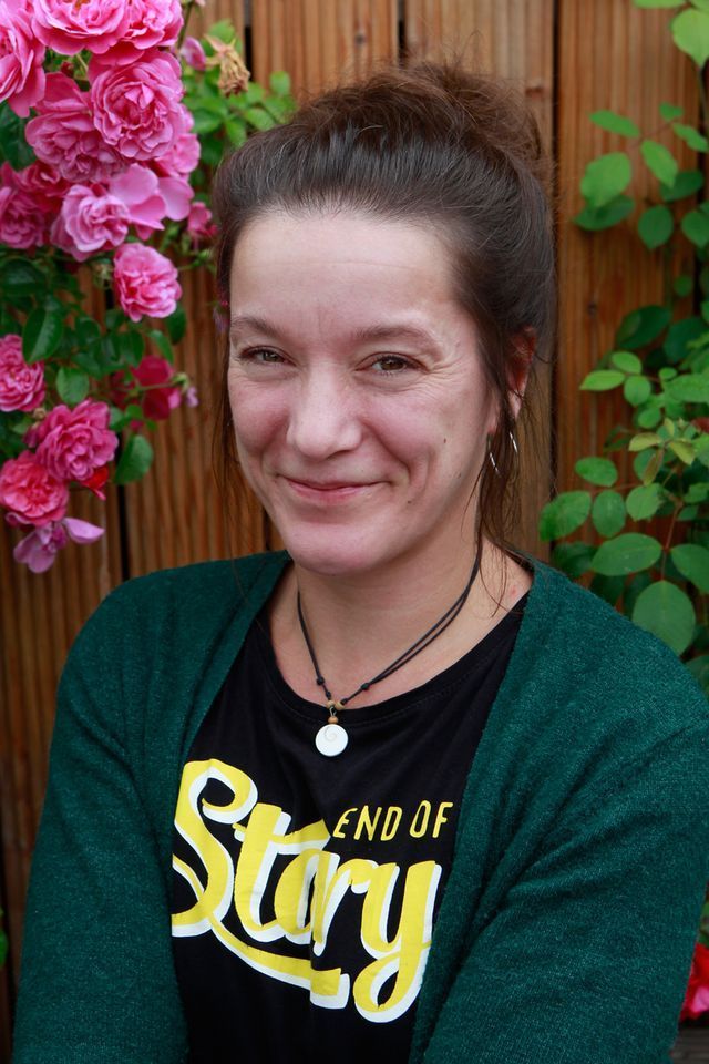 Frau in grüner Strickjacke lächelt, Blumen im Hintergrund.