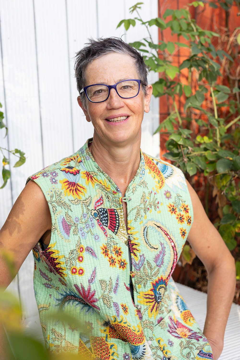 Frau mit Brille lächelt und trägt eine ärmellose, gemusterte Weste. Im Freien in der Nähe eines weißen Zauns und grüner Pflanzen.