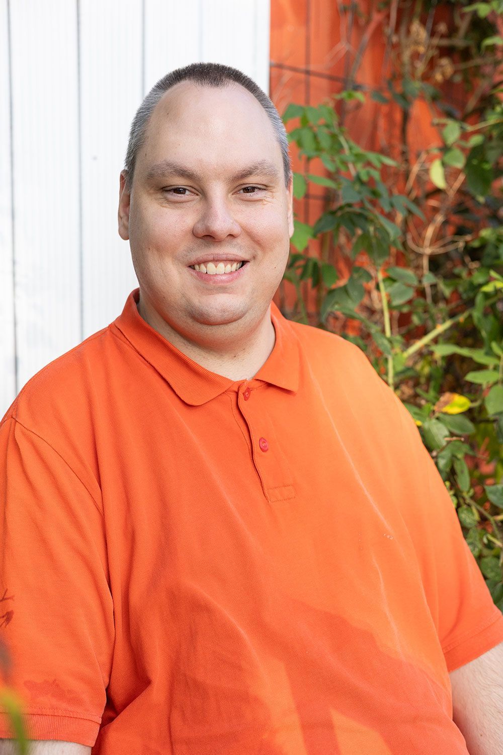 Ein Mann in einem orangefarbenen Poloshirt lächelt im Freien vor einer weiß-orangen Wand mit grünem Laub.