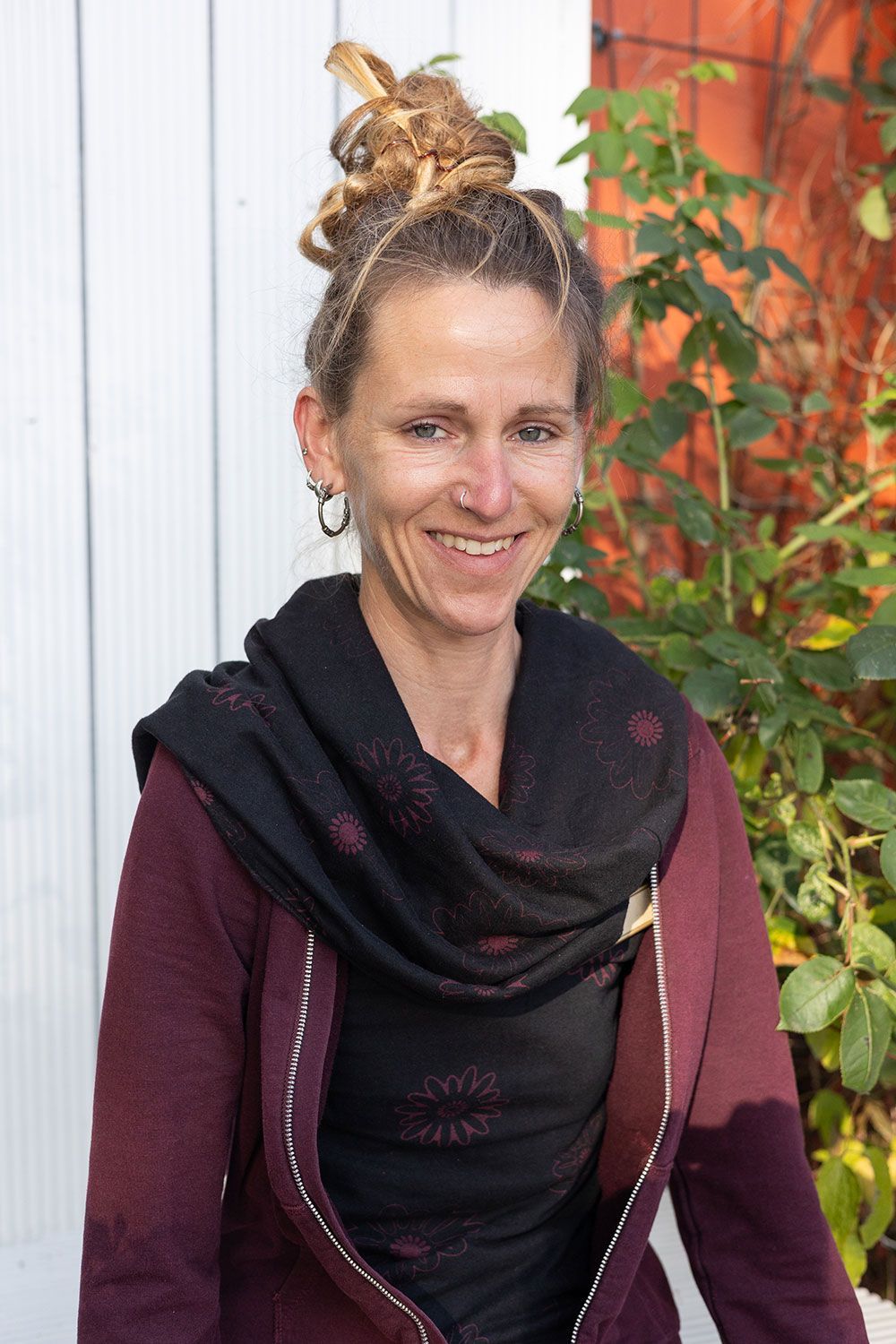 Frau mit Dutt, lächelnd, mit dunklem Schal und kastanienbrauner Jacke, steht vor einer weißen Wand und viel Grün.