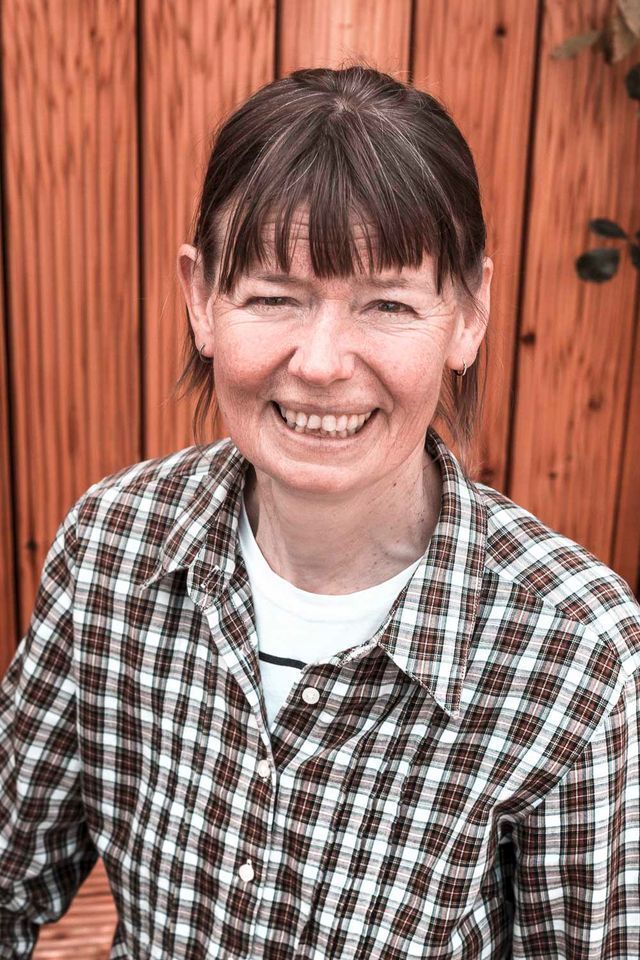 Lächelnde Frau in kariertem und weißem Hemd vor einem Holzzaun.