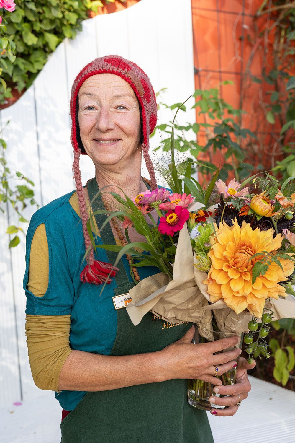 Frau mit rotem Hut, buntem Blumenstrauß in der Hand, lächelnd. An ein weißes Tor gelehnt.