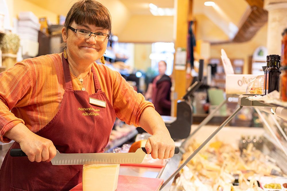Frau schneidet Käse in einem Feinkostladen und lächelt. Burgunderfarbene Schürze, orangefarbenes Hemd, Brille. Ladeneinrichtung mit Glasvitrine.