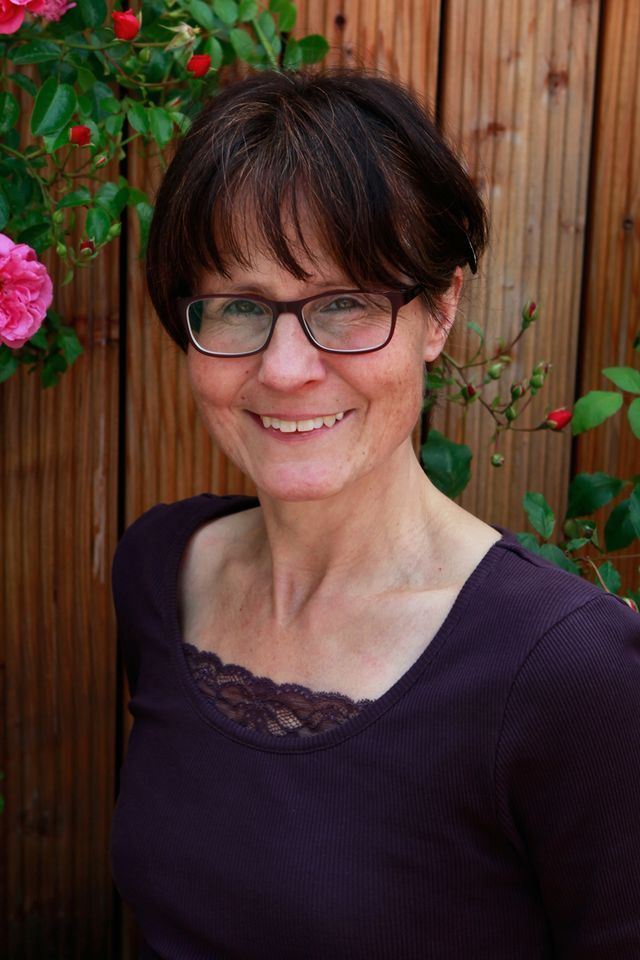 Frau mit Brille lächelt vor einem Holzzaun mit rosa Rosen.