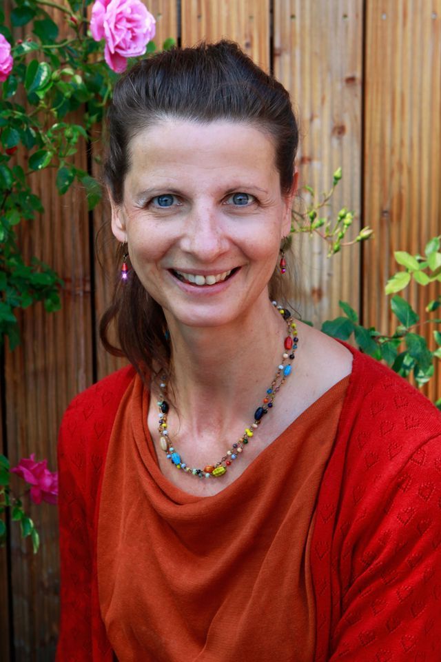 Lächelnde Frau mit Halskette und rotem Oberteil vor einem Holzzaun mit rosa Rosen.