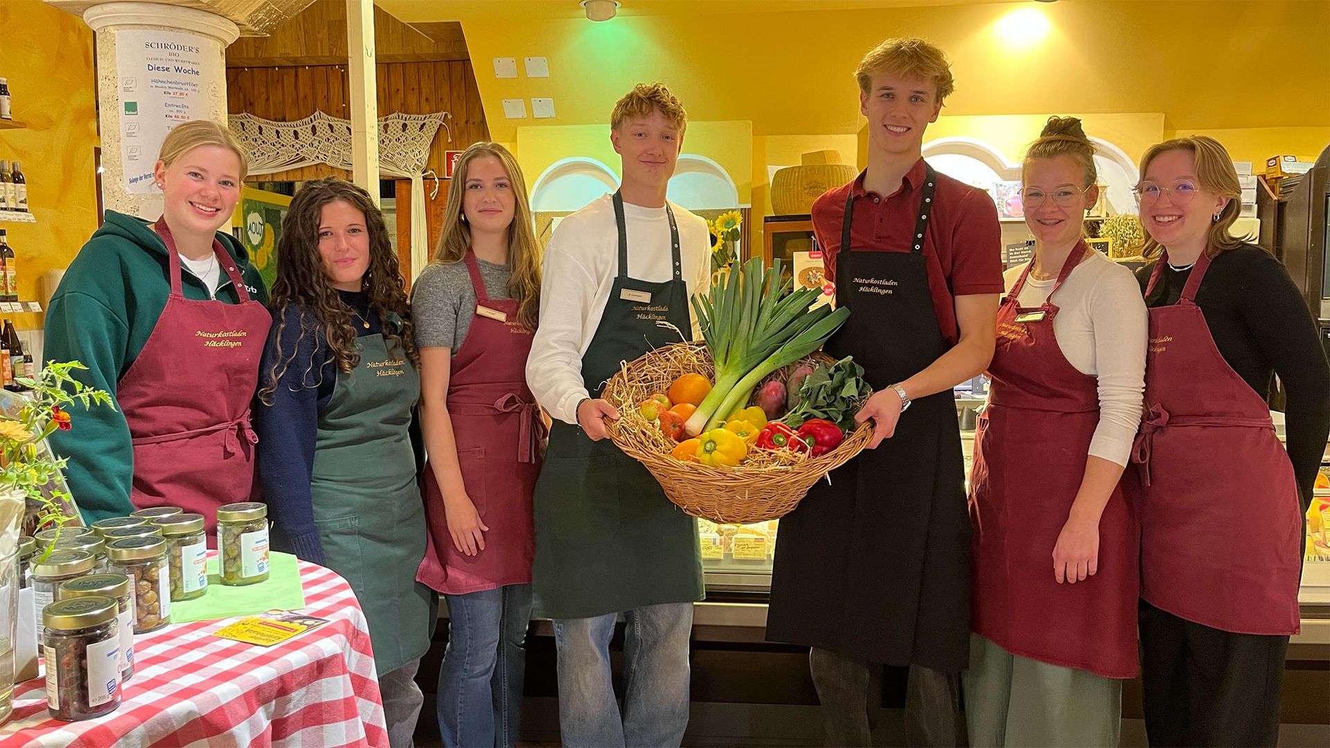 Gruppe von Menschen in Schürzen mit einem Korb voller Gemüse auf einem hell erleuchteten Markt.