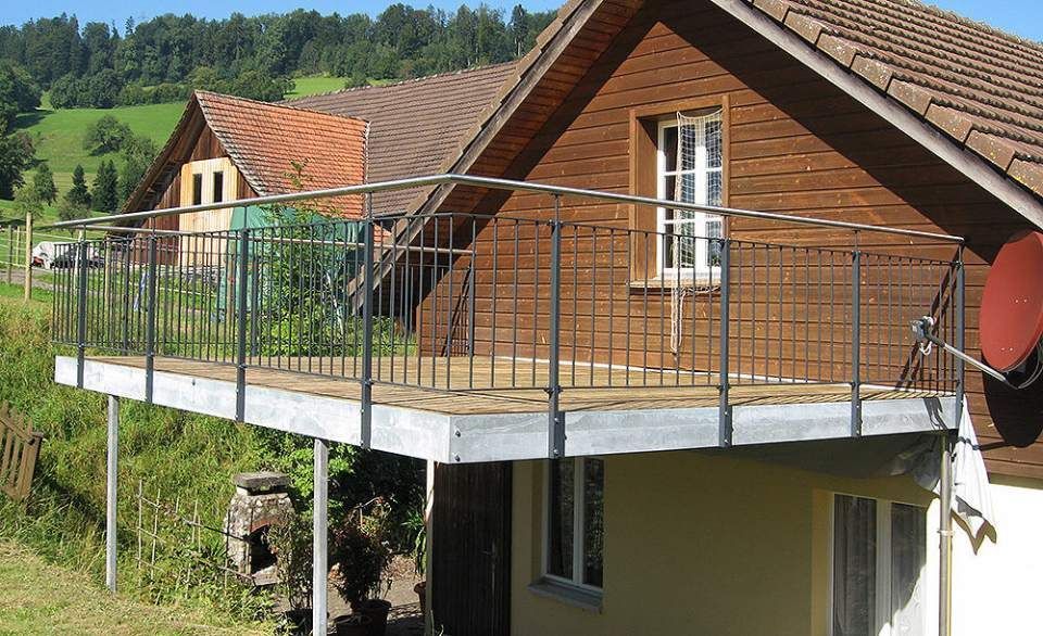 Balkon mit Metallgeländer an einem holzverkleideten Haus, das an einem grasbewachsenen Hang liegt.
