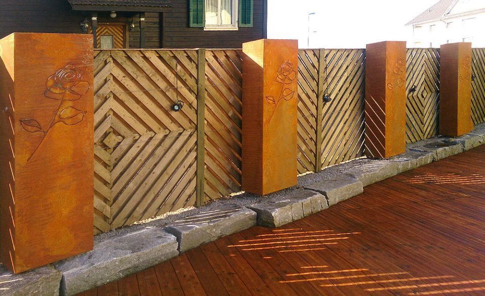 Ein Holzzaun mit rostigen Metallpfosten auf einem Steinsockel bildet eine dekorative Abgrenzung.
