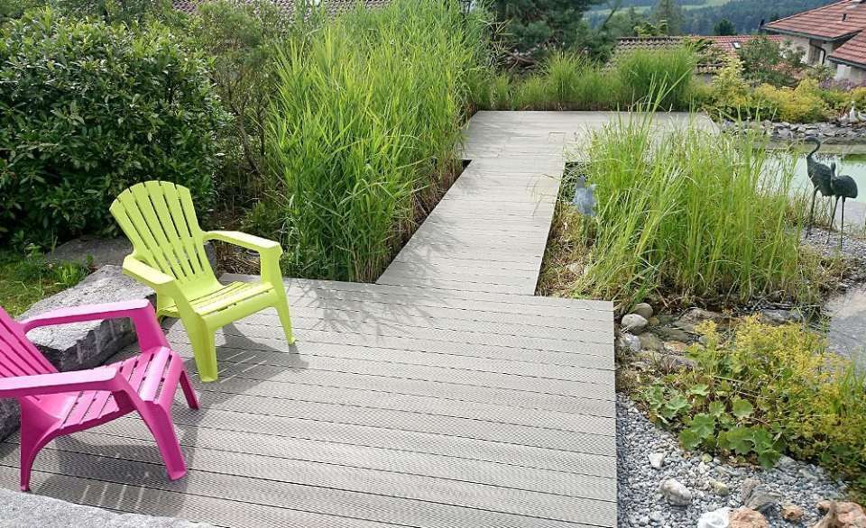 Holzterrasse mit grünen und rosa Stühlen, angrenzend an hohes Gras und einen Teich.