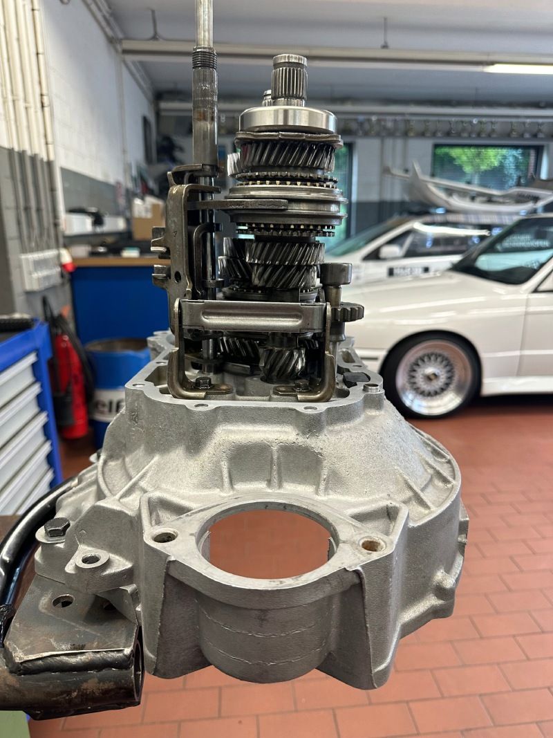 Freiliegendes Autogetriebe auf einer Werkbank in einer Autowerkstatt, im Hintergrund ein weißes Auto.