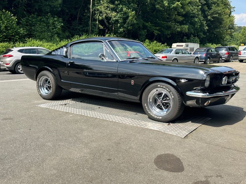 Schwarzer klassischer Ford Mustang, geparkt auf einem Parkplatz in der Nähe von Bäumen und anderen Autos.