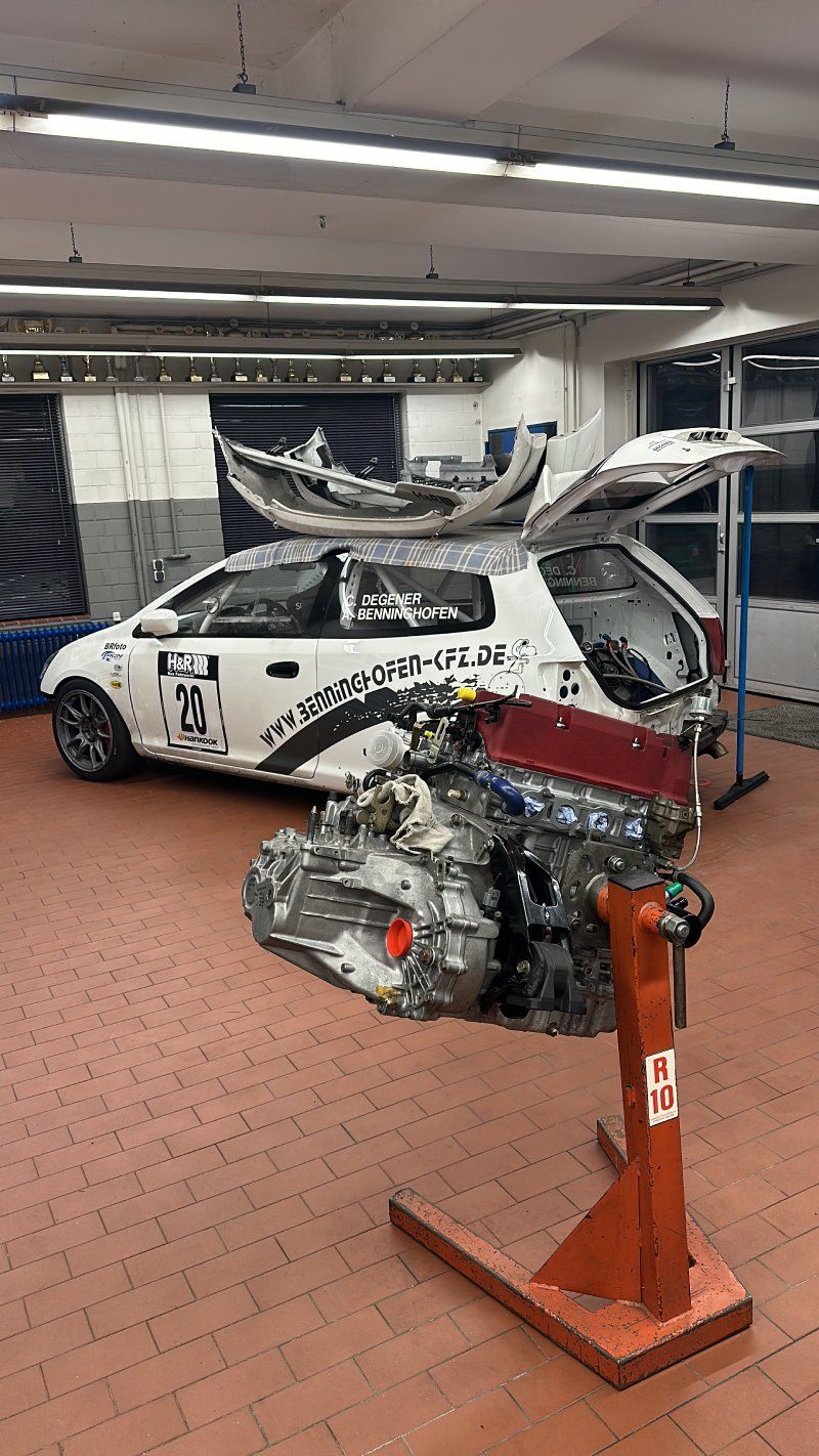 Beschädigtes weißes Auto mit freiliegendem Motor in einer Werkstatt, montiert auf einem orangefarbenen Ständer im Vordergrund