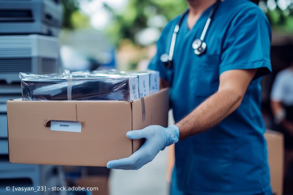 Medizinische Fachkraft in OP-Kleidung und Handschuhen trägt Kisten nach draußen.