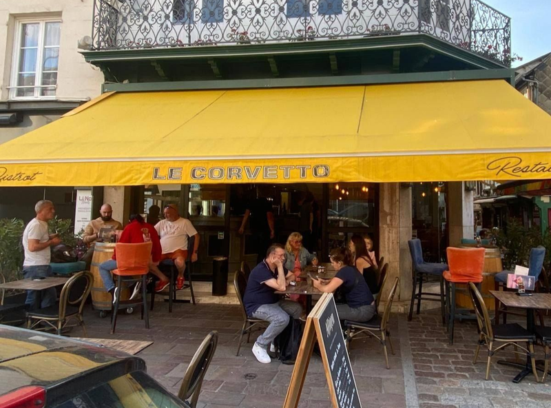 Un auvent jaune surplombe un bistro nommé Le Corvetto. Des gens sont assis à des tables en terrasse, certains en train de boire.