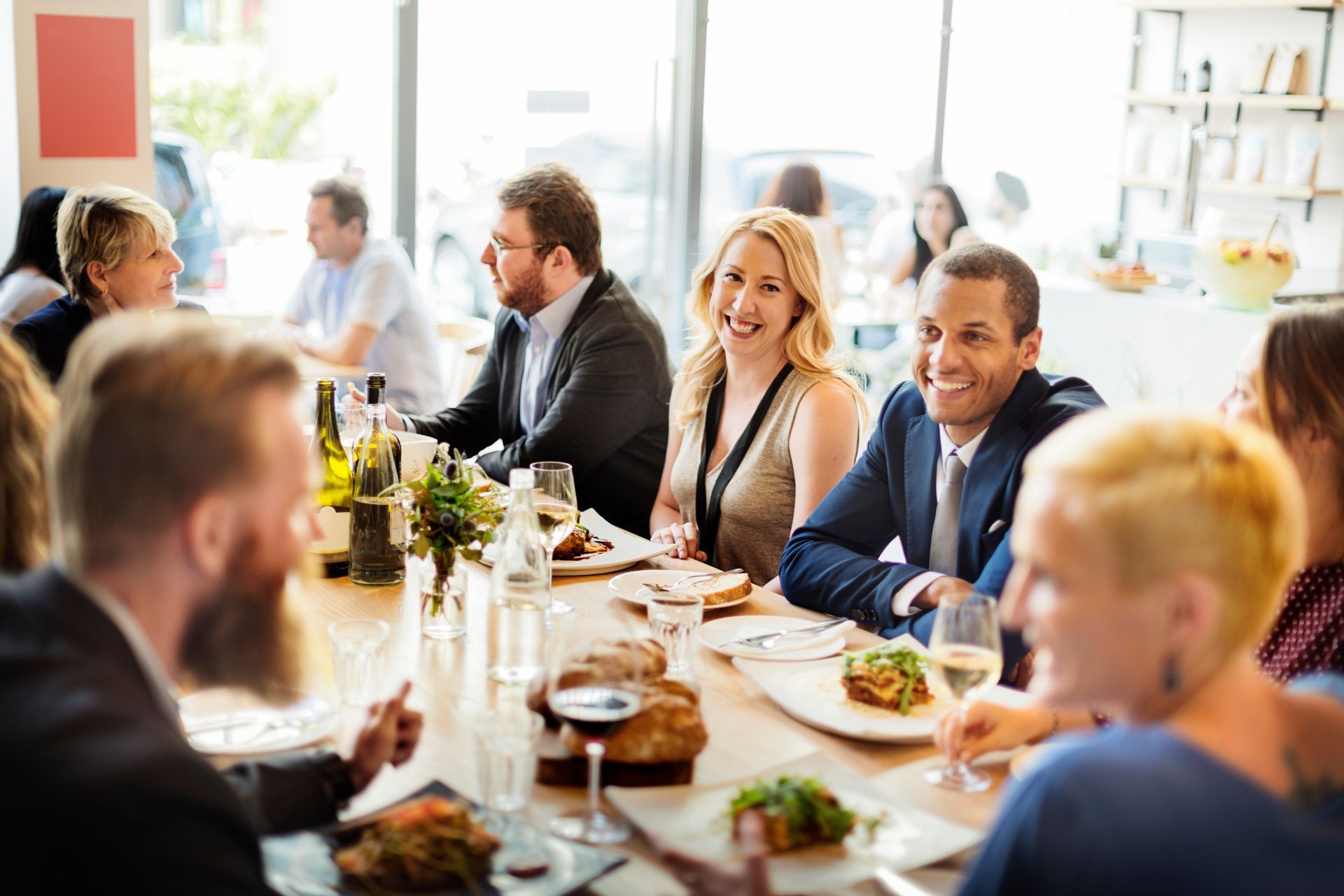 Des gens sourient et mangent à une longue table dans un restaurant. La lumière du soleil entre par une fenêtre.