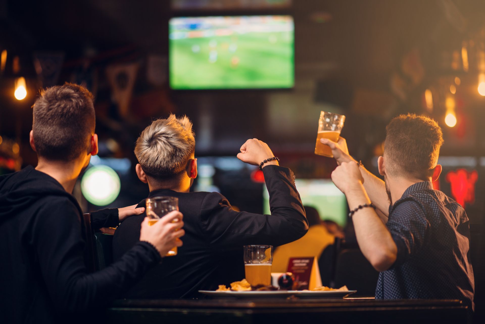 Trois personnes dans un bar, applaudissant en regardant un match de sport à la télévision, un verre de bière à la main.