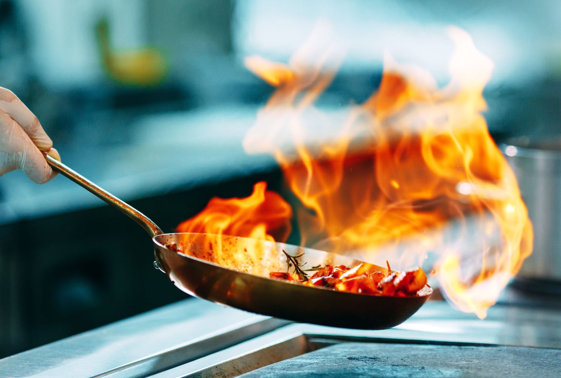 Des flammes jaillissent d'une poêle tenue à la main ; des aliments cuisent dans une cuisine.