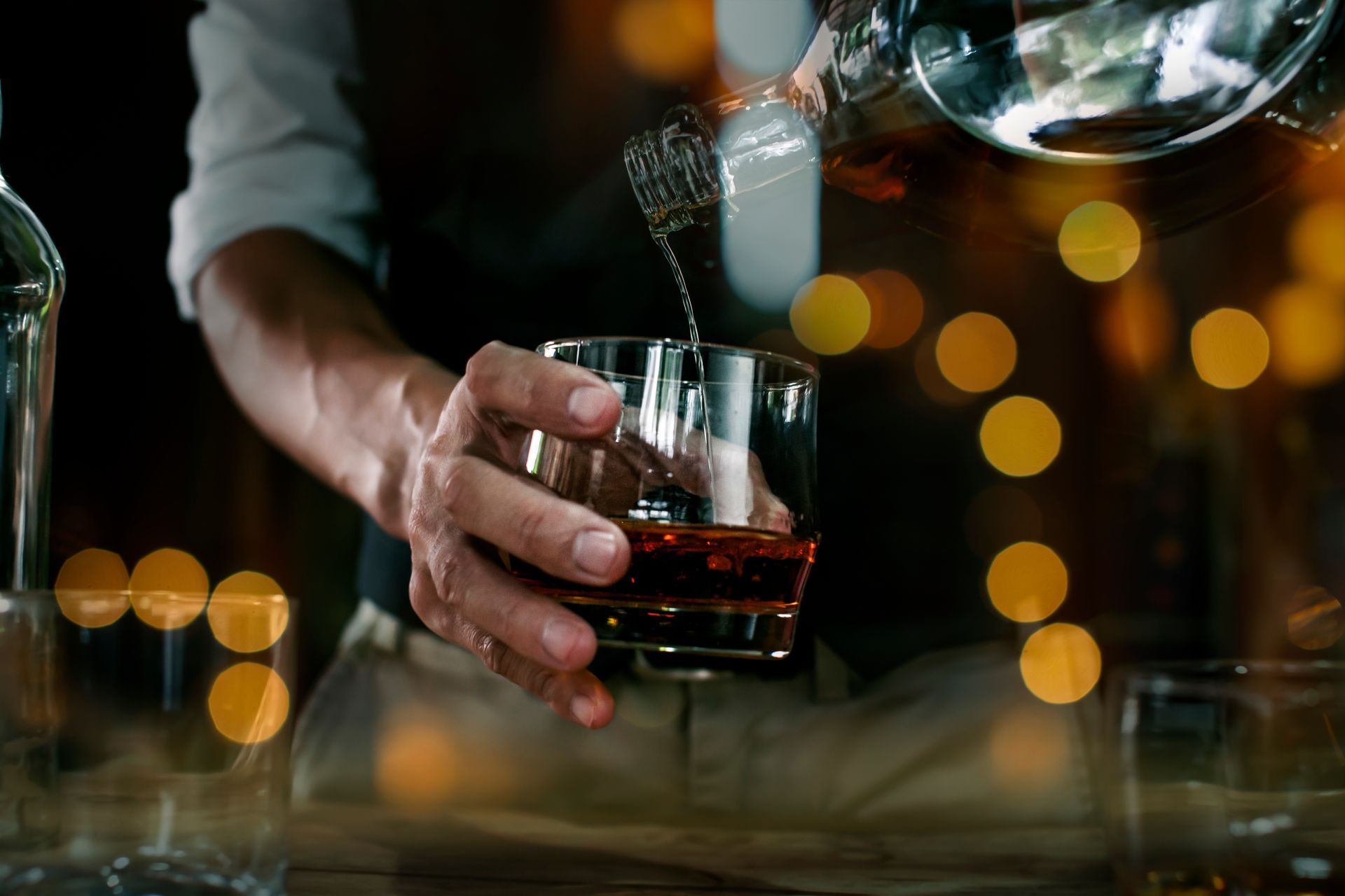 Un barman verse du whisky dans un verre dans un bar, avec des lumières bokeh en arrière-plan.