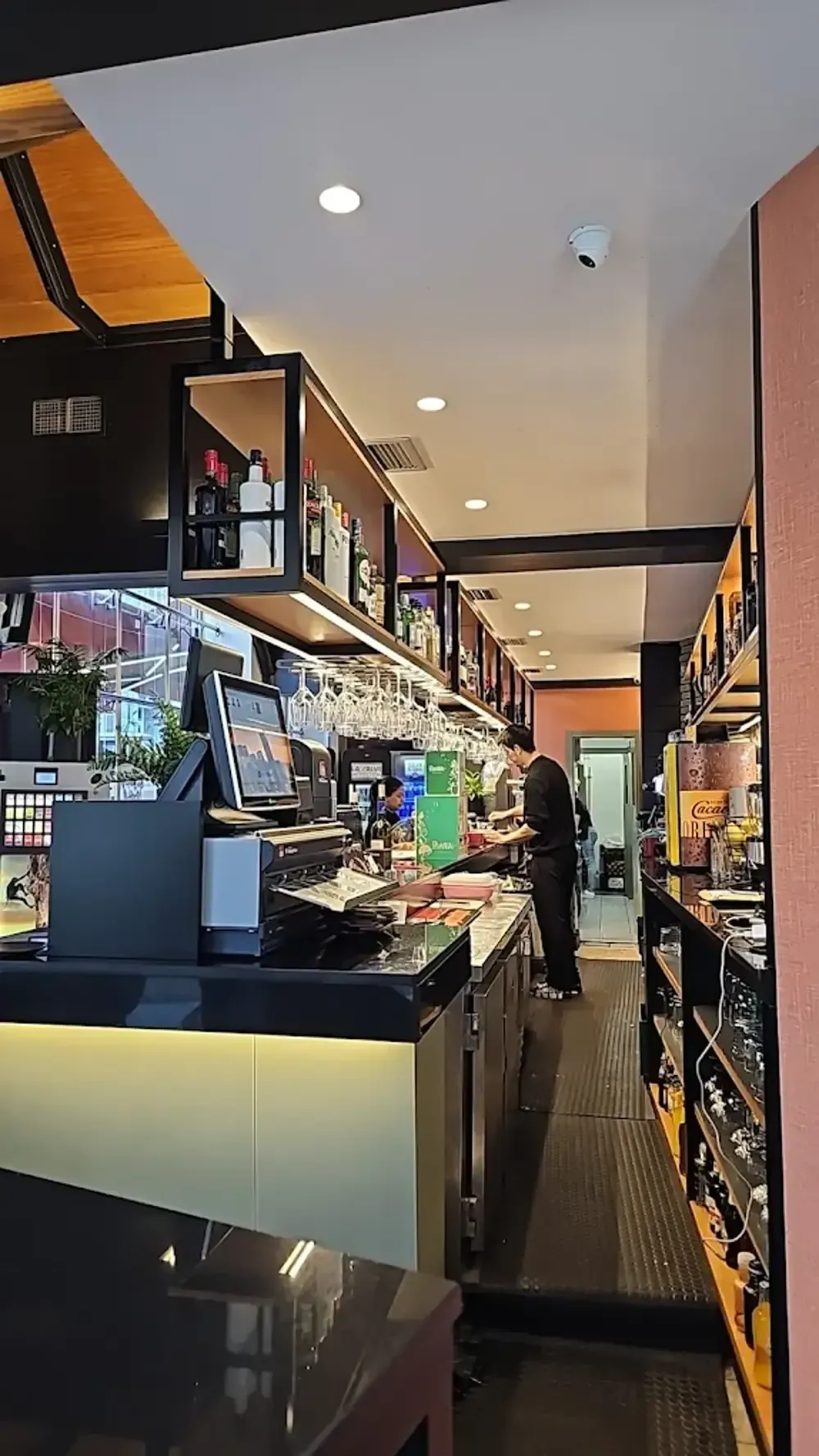 Vista interior de un bar con un camarero. Detalles en negro y dorado, estantes llenos de botellas e iluminación cenital.