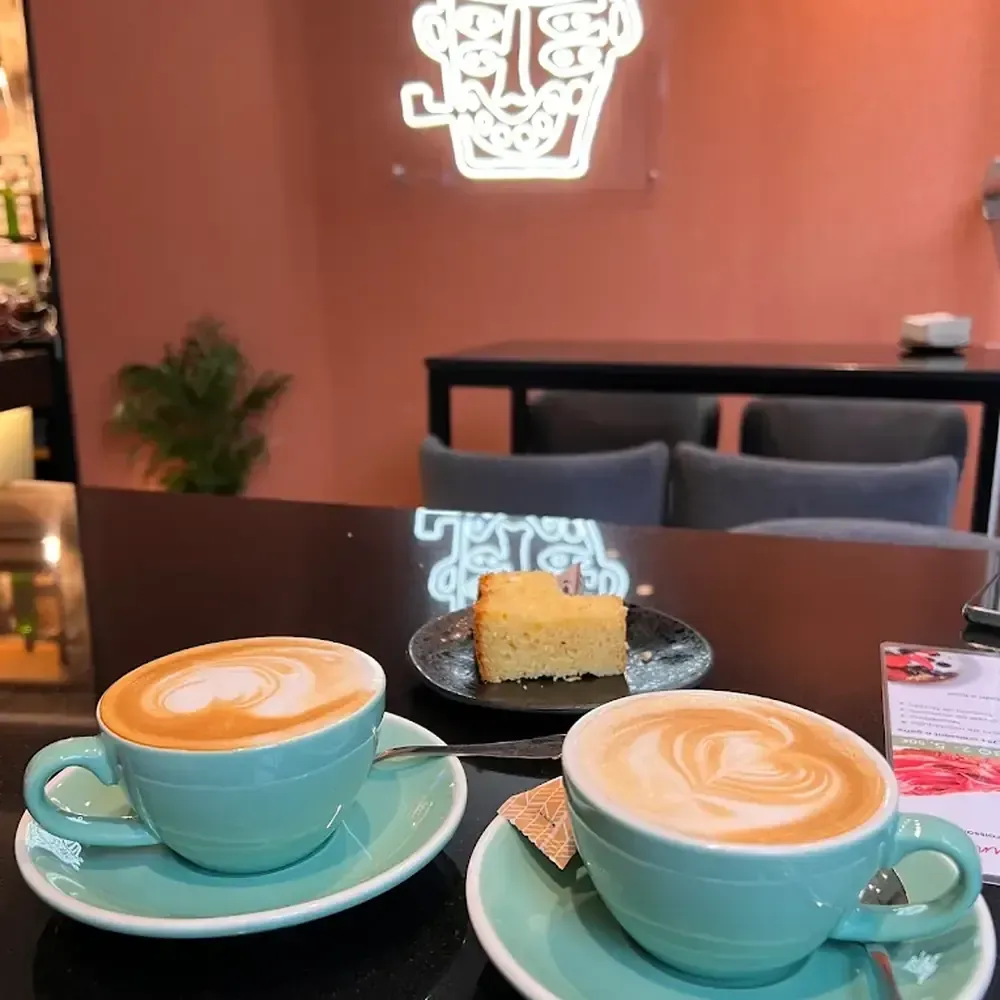 Dos cafés con leche en tazas color verde azulado sobre platillos con una rebanada de pastel en un plato negro.