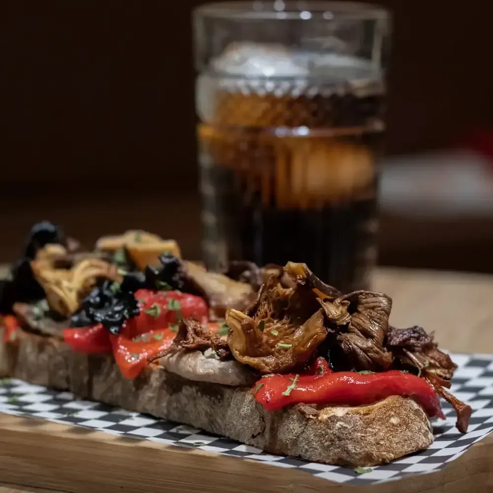 Pan tostado cubierto con champiñones, pimientos y hierbas, con un vaso de líquido oscuro de fondo.