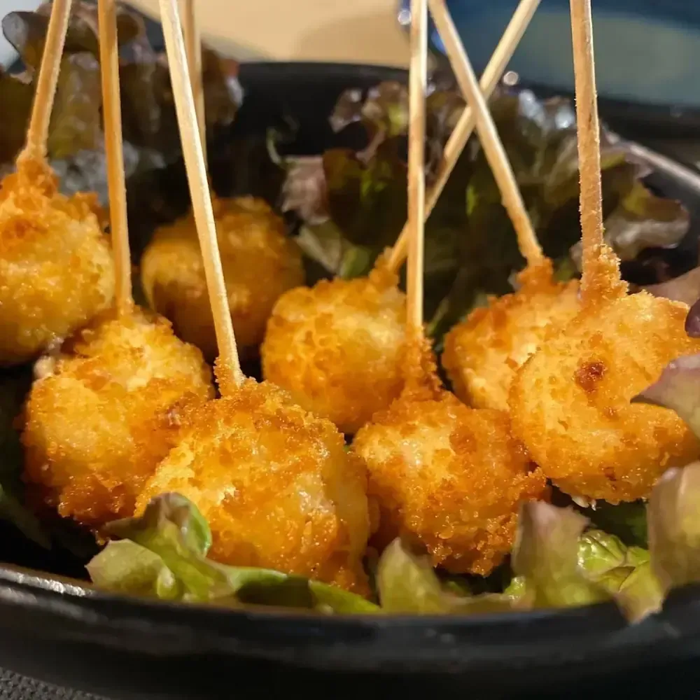 Comida frita y dorada en brochetas, dispuesta sobre lechuga en un recipiente oscuro.